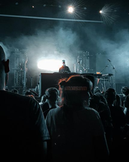Concert crowd facing a stage with a DJ. Stage lit with spotlights and smoke.