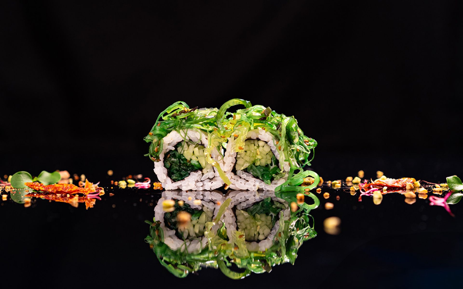 Rotoli di sushi con alghe, verdure e riflessi, su sfondo nero e guarnizioni colorate.