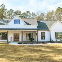 A white house with a porch and a gray roof is sitting on top of a lush green field.