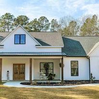 A white house with a porch and a gray roof is sitting on top of a lush green field.