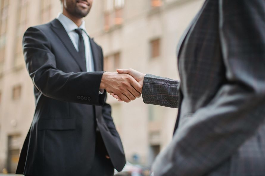 Two people in professional suits shaking hands in an outdoor setting.