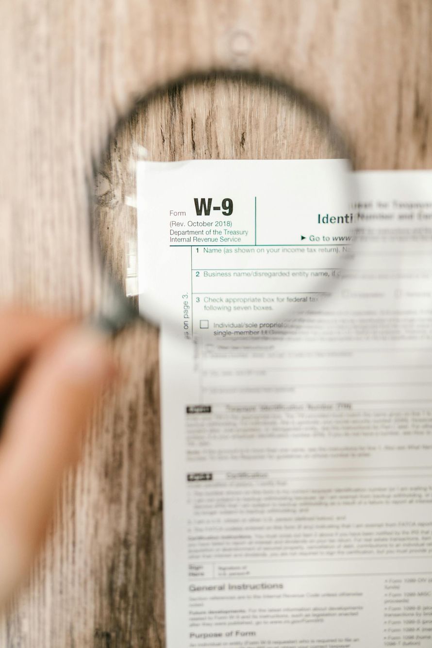 A magnifying glass held over a W-9 tax form on a wooden surface.