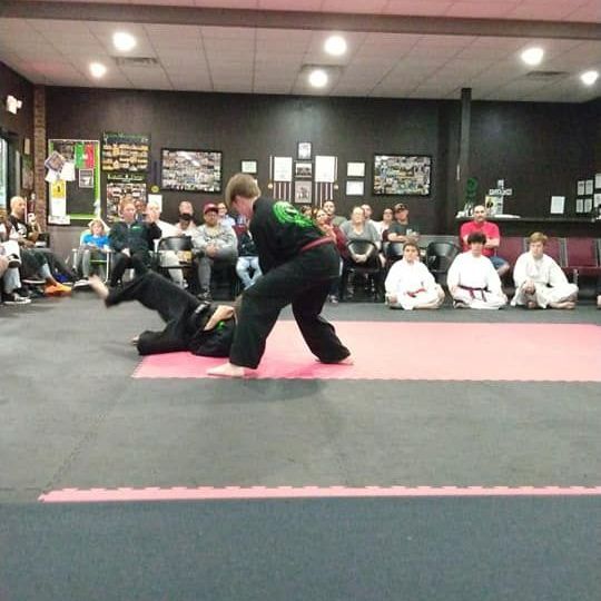 Two martial artists sparring on a mat in a dojo. One is falling while the other stands.