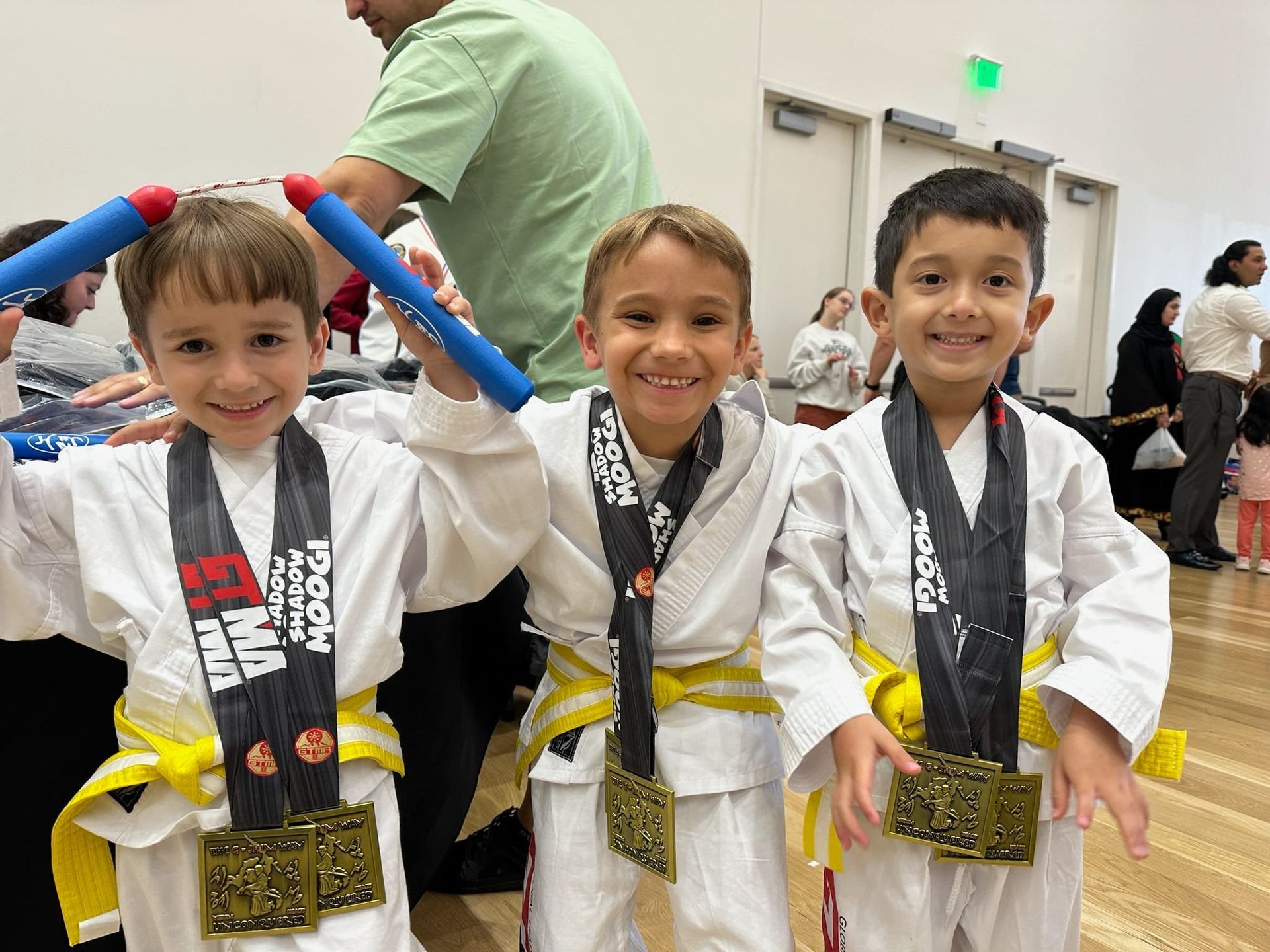 Three young boys in martial arts uniforms with medals, smiling, inside a building.