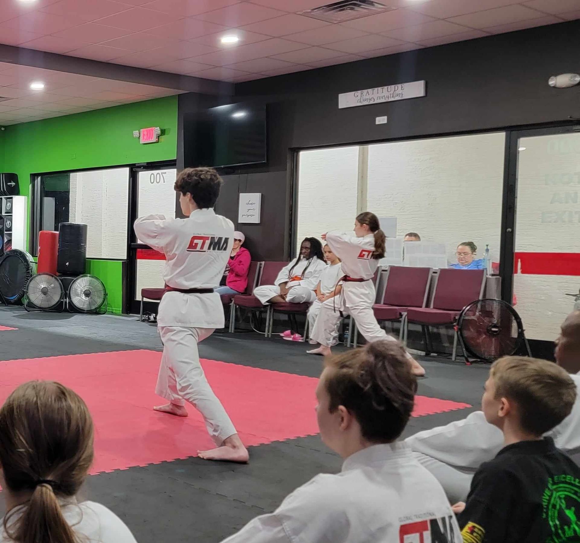 Martial arts class: Two students practice moves, watched by peers. Red mat, white uniforms, green and black walls.