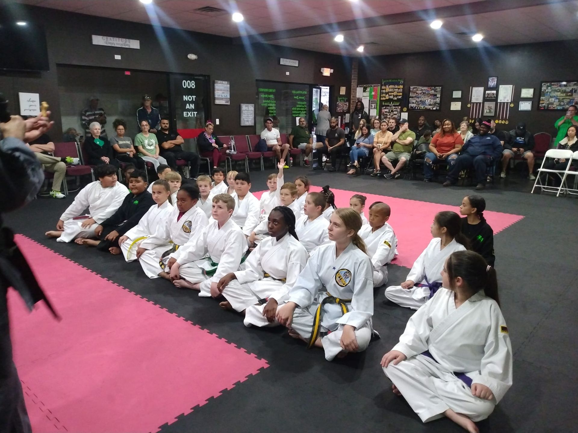 Children in karate uniforms sitting, facing away from audience in a dojo.