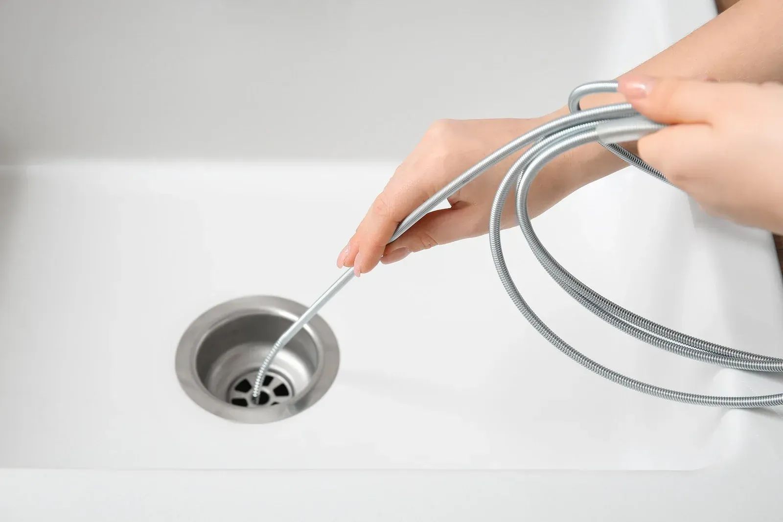 Hands using a drain snake to unclog a white sink drain.