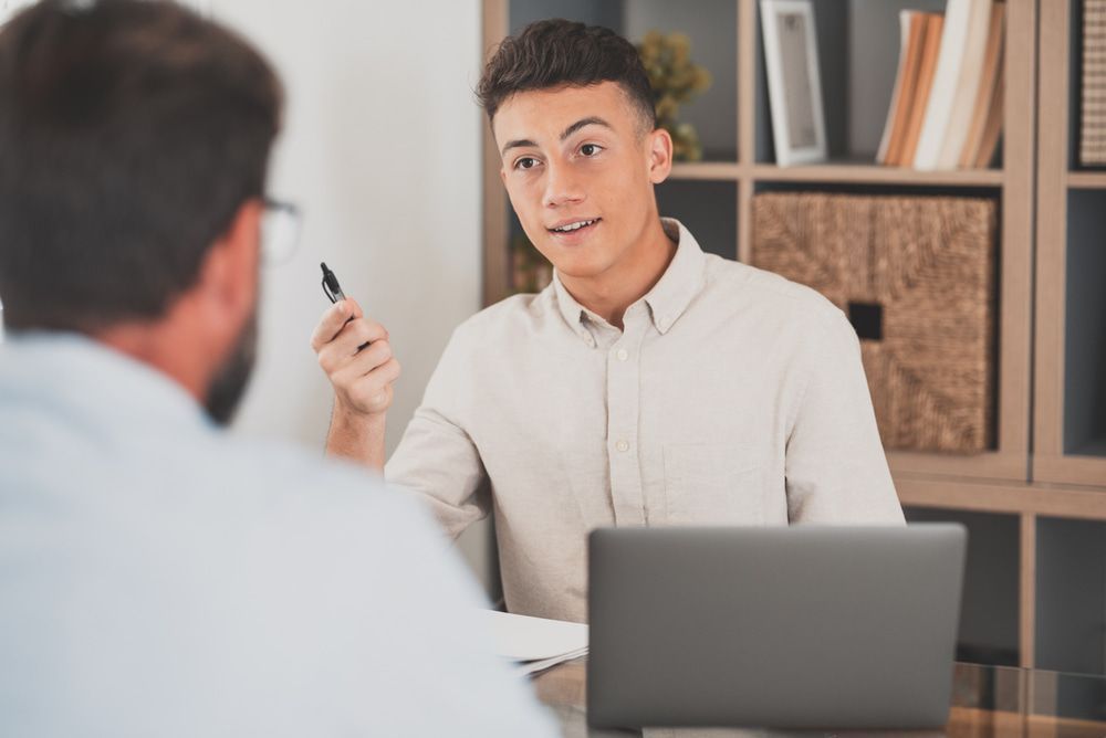 A Man Is Sitting At A Table With A Laptop And Talking To Another Man — Black Label I.T. in Garbutt, QLD