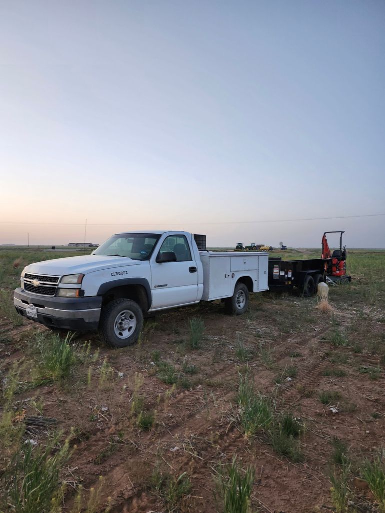 White work truck with utility bed parked in a field, under a clear sky. A trailer with equipment is attached.