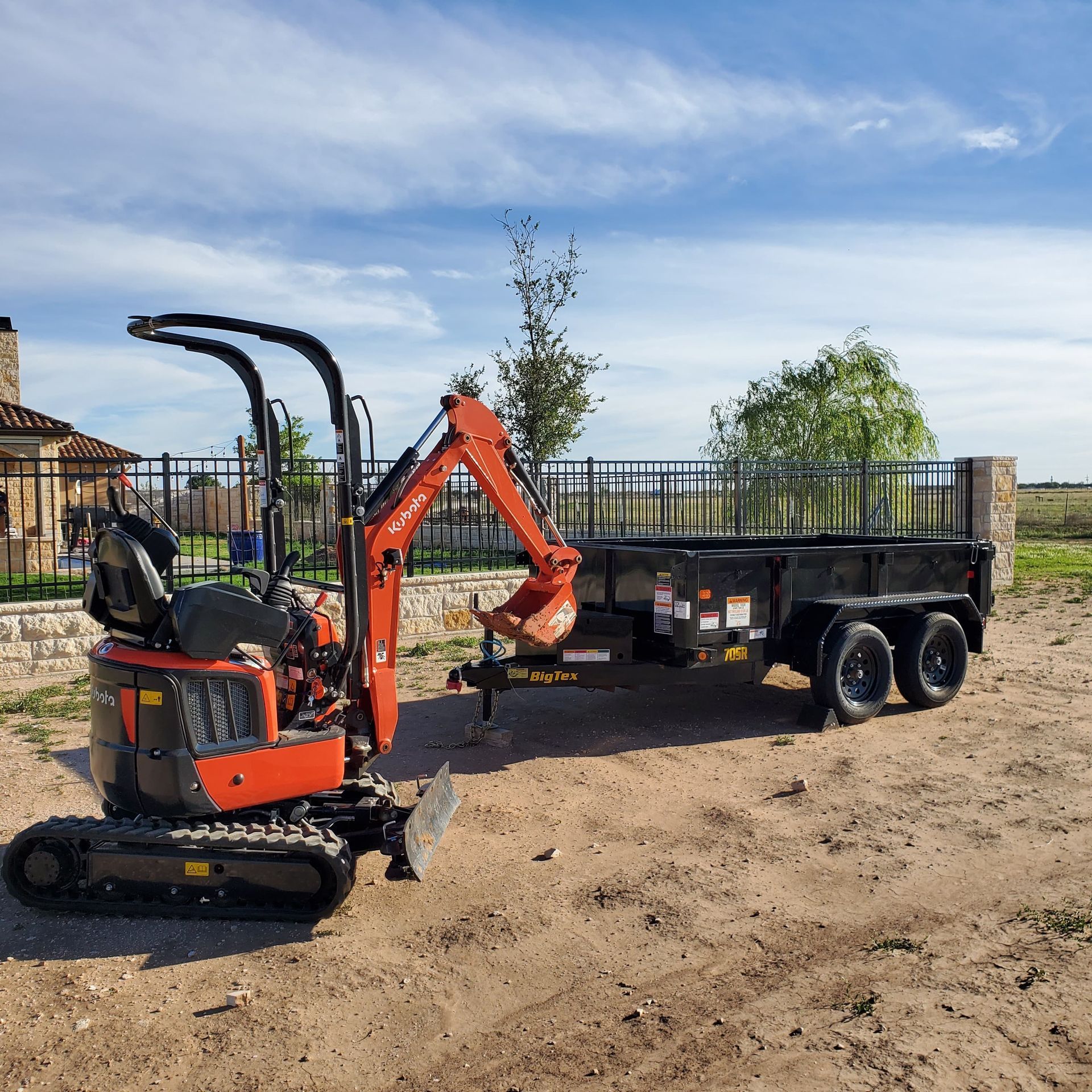 Orange mini excavator next to a black trailer on a dirt surface under a blue sky.