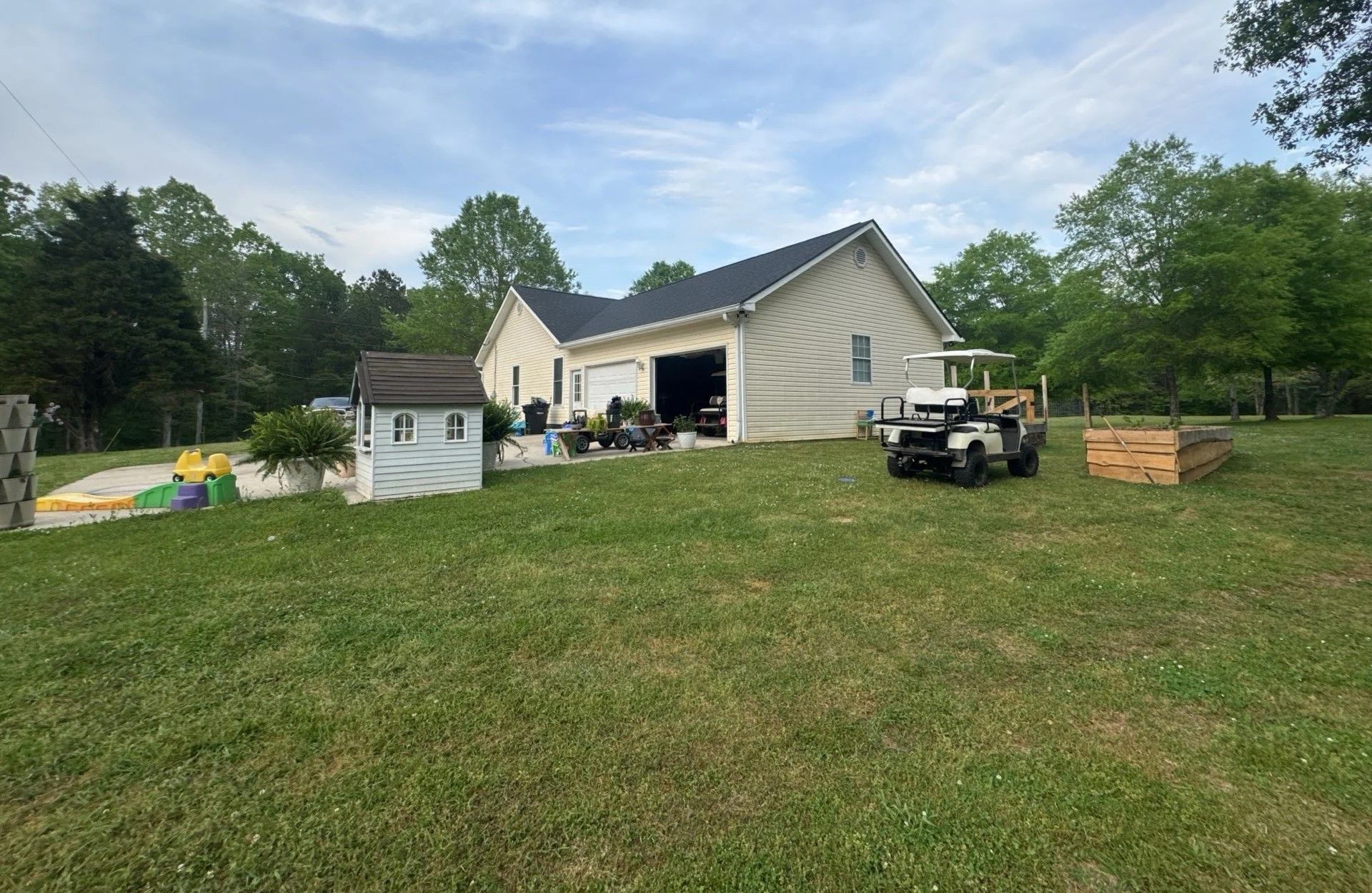 Backyard with house, playhouse, cart, and garden bed on a grassy lawn under a cloudy sky.