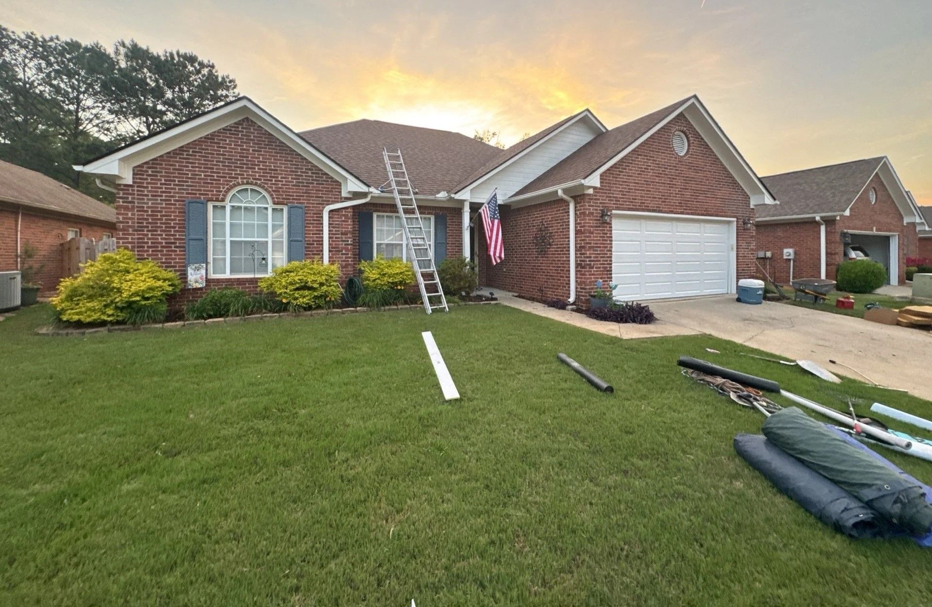 Brick house with ladder on roof, lawn, and construction materials on the ground; sunset in the background.
