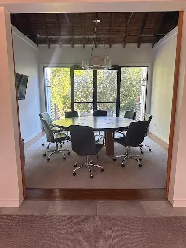 Meeting room with round table and chairs, large window overlooking trees, neutral colors.