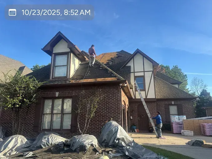 Two workers on roof replacing shingles, brick house, sunny day, ladder.