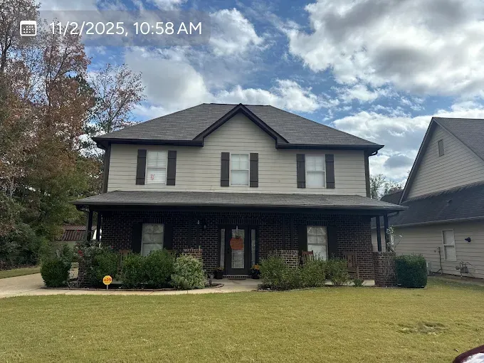 Two-story house with tan siding, brown shutters, a brick porch, and a brown roof. Overcast sky.