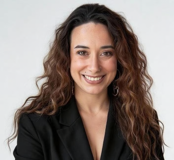 Woman with long, wavy brown hair smiles broadly, wearing a black blazer against a white backdrop.
