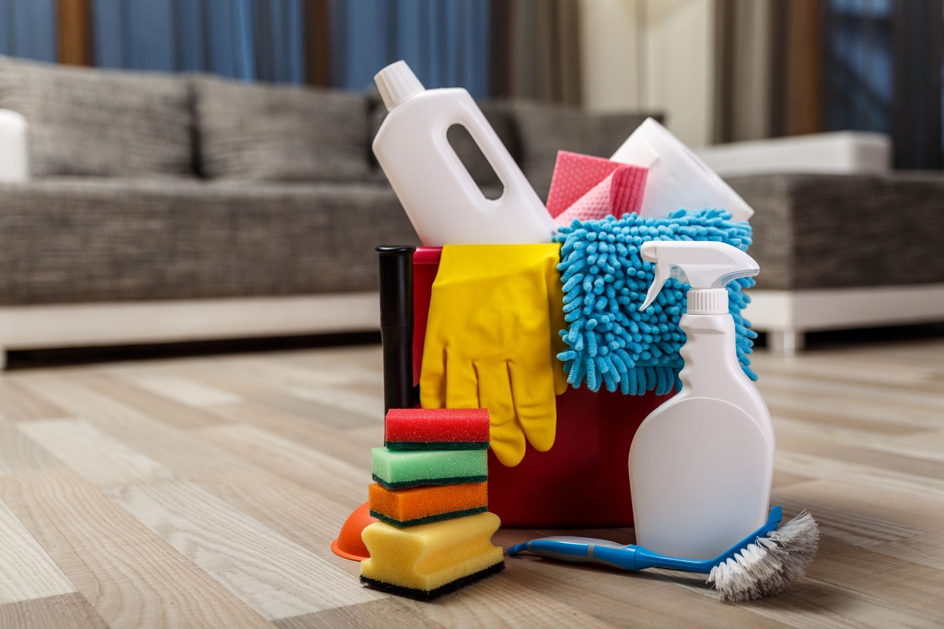 Cleaning supplies in a red bucket on a wood floor: sponges, spray bottle, gloves, cloths, and a brush, near a couch.
