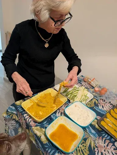 Una persona impana delle fette di zucchina in un piatto giallo, con ciotole contenenti uova e farina accanto, su un tavolo apparecchiato con una tovaglia a fiori.