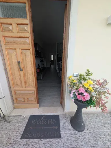 Una porta di legno aperta conduce in una casa con uno zerbino di benvenuto e accanto una fioriera alta piena di fiori colorati.