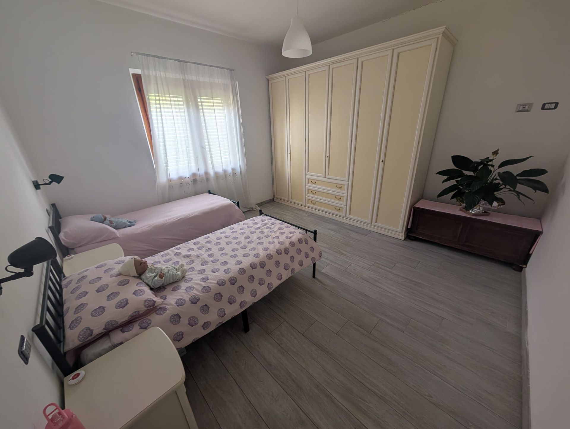 Una camera da letto con due letti singoli, un grande armadio color crema, una piccola panca con una pianta e pavimento in legno grigio chiaro.