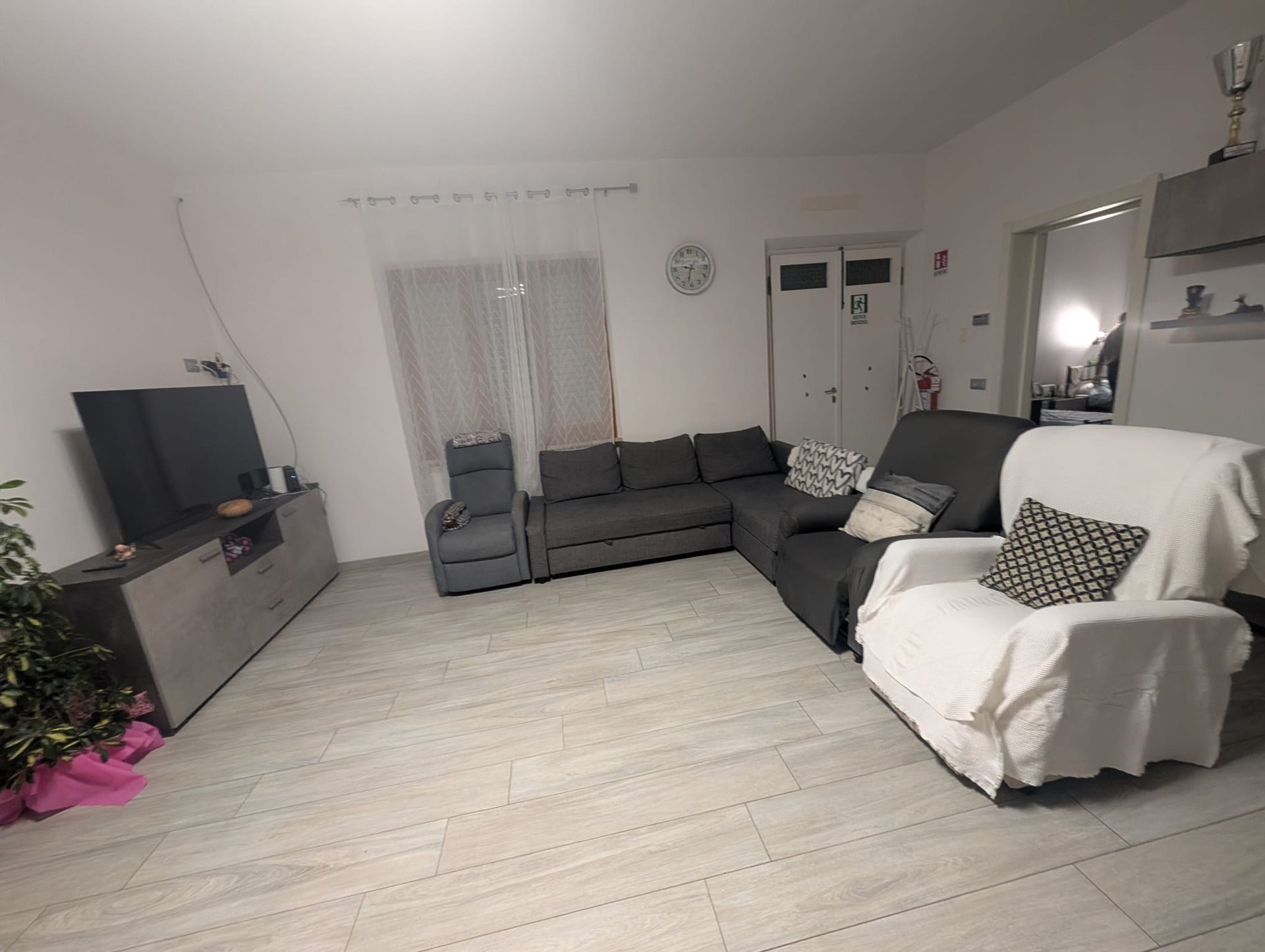 Un soggiorno con un grande divano angolare grigio, una poltrona bianca, un mobile porta TV e un pavimento in legno chiaro.