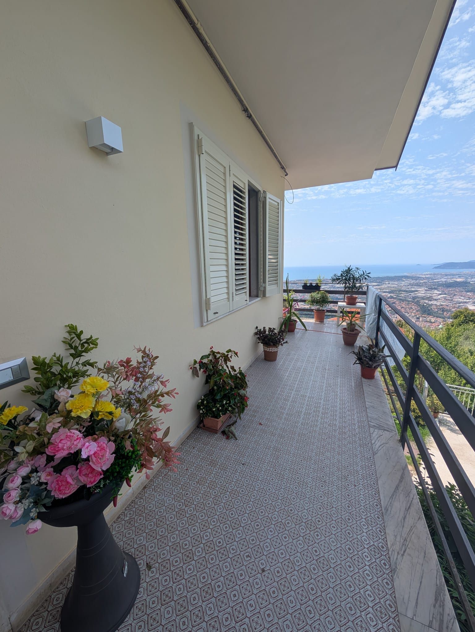 Un balcone con piante in vaso, persiane decorative alla parete e una vista che spazia sulla città e sul mare.