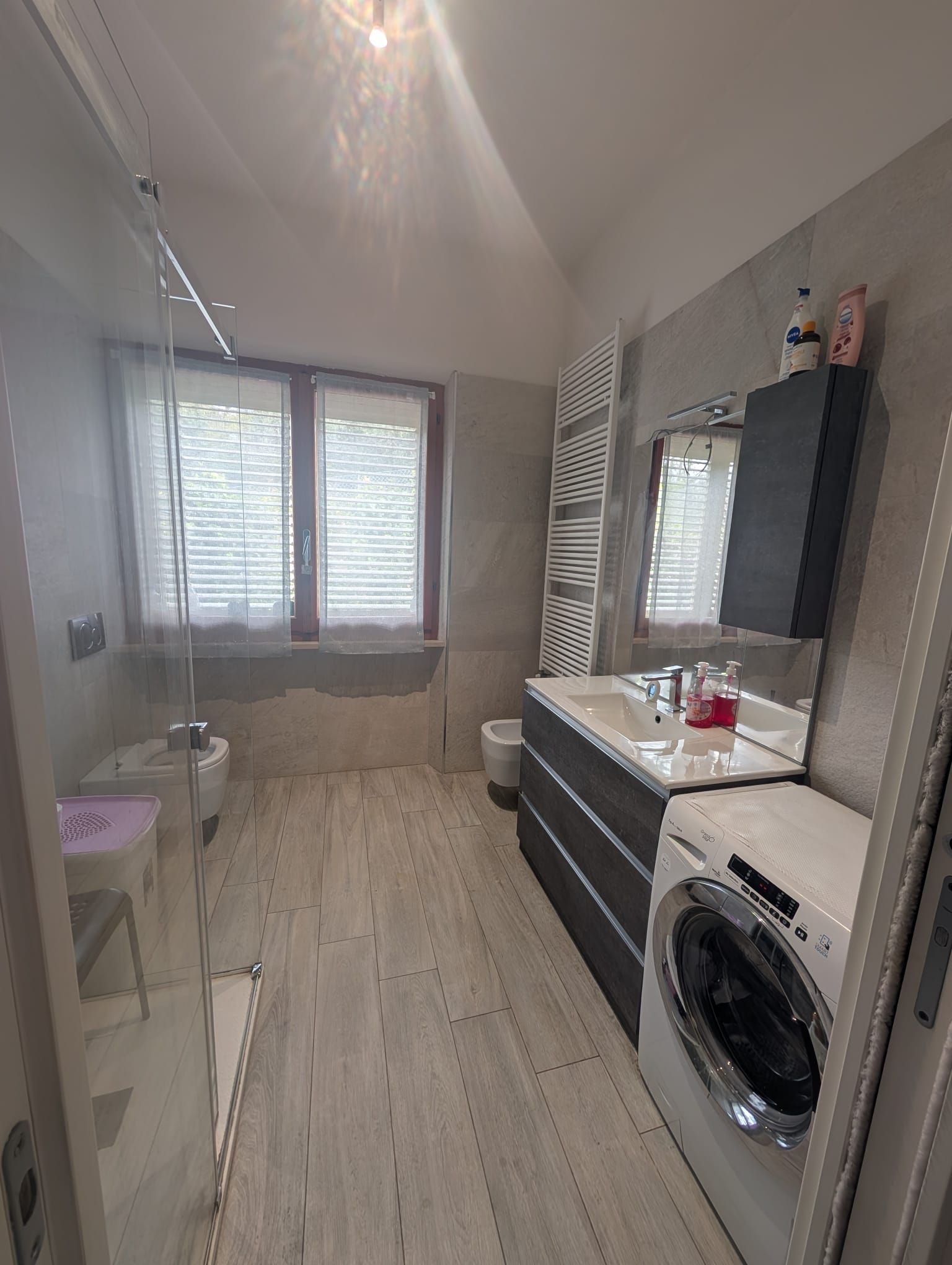 Un bagno moderno con mobile lavabo bianco, lavatrice a carica frontale, bidet, cabina doccia e finestre con tende.