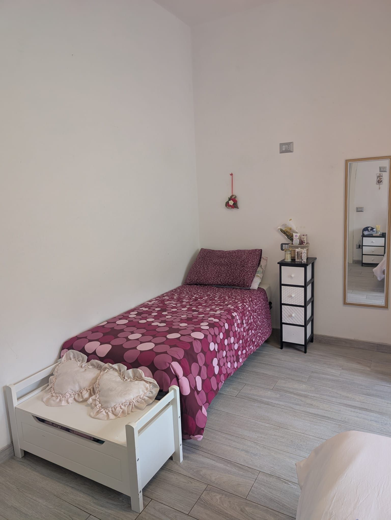 La camera da letto è arredata con un letto singolo con una trapunta viola a motivi geometrici, una panca bianca con cuscini a forma di cuore e uno specchio.