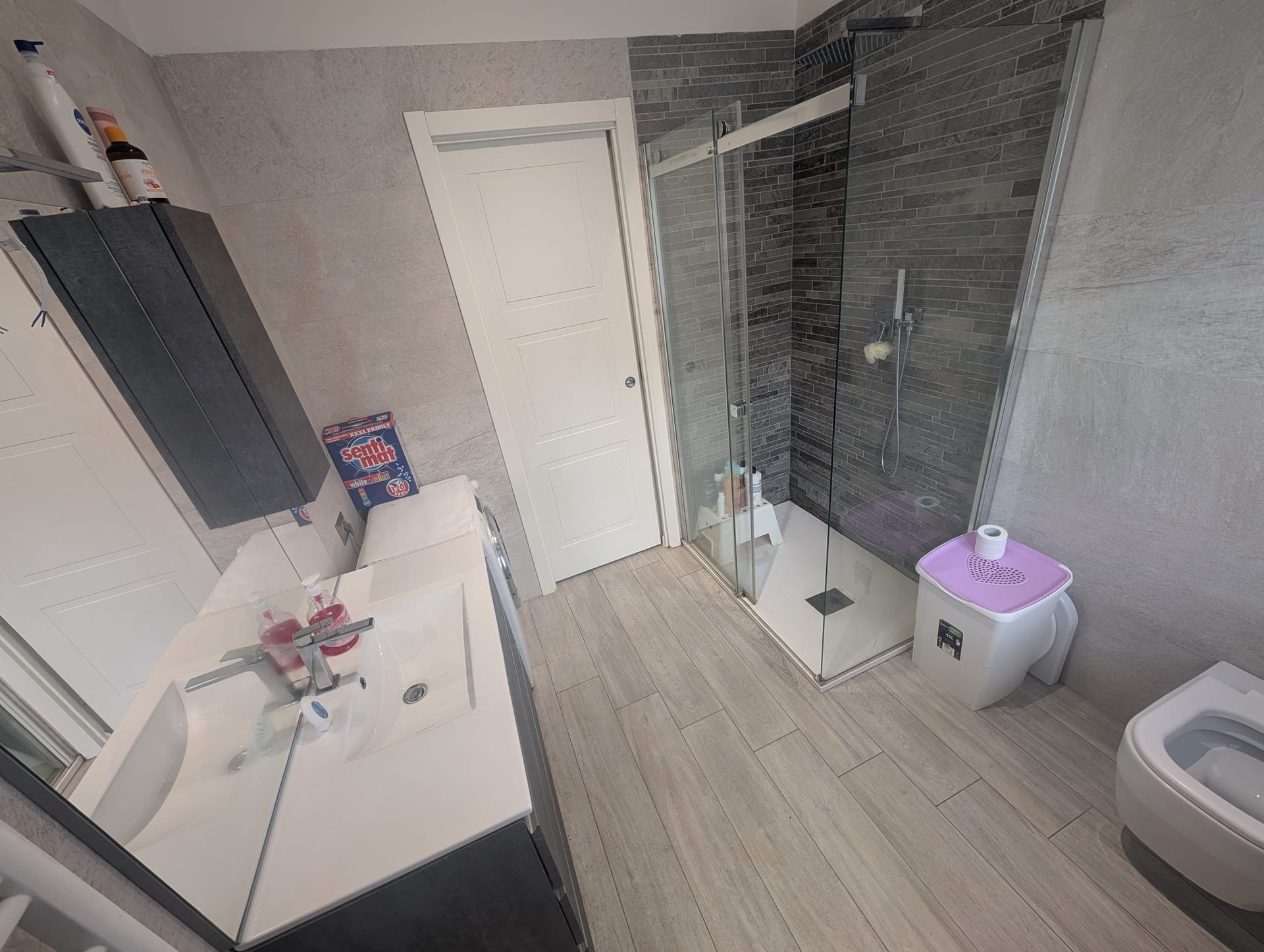 Un bagno con mobile lavabo bianco, cabina doccia con pareti rivestite di piastrelle grigie, WC bianco con copriwater rosa e bidet.