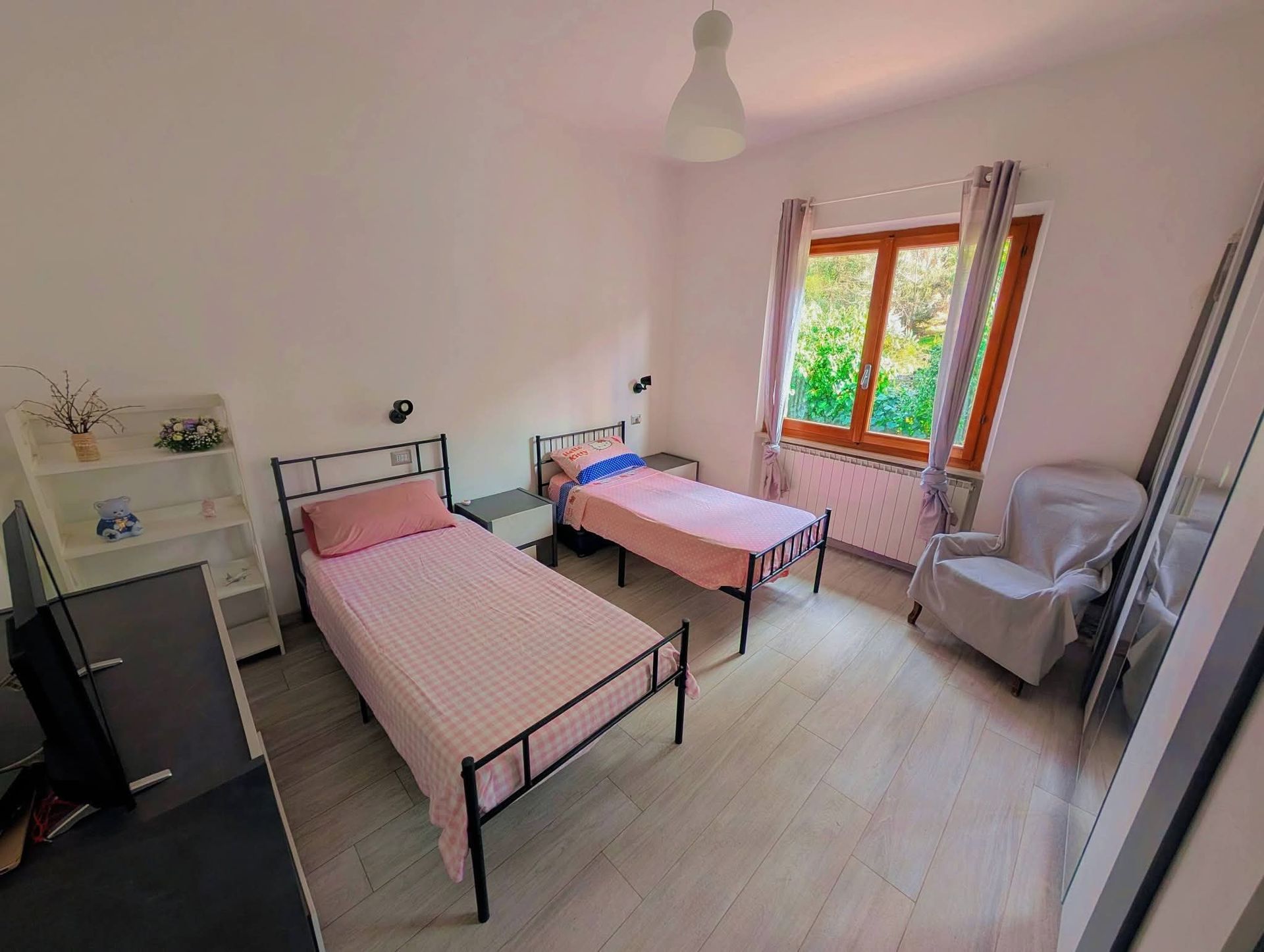 Una camera da letto luminosa e arredata in modo essenziale, con due letti singoli con coperte rosa, una mensola bianca e una finestra con tende.