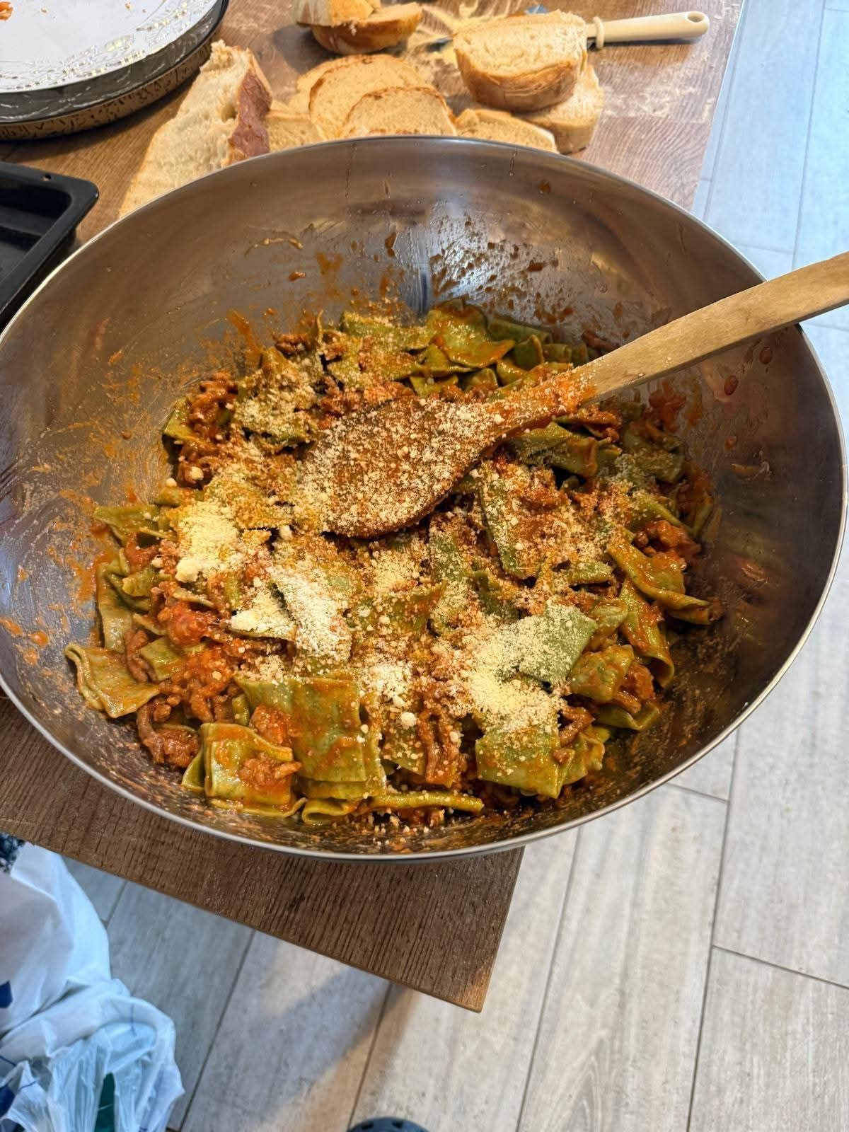Una ciotola di metallo piena di pasta verde condita con salsa rossa, cosparsa di formaggio e mescolata con un cucchiaio di legno.