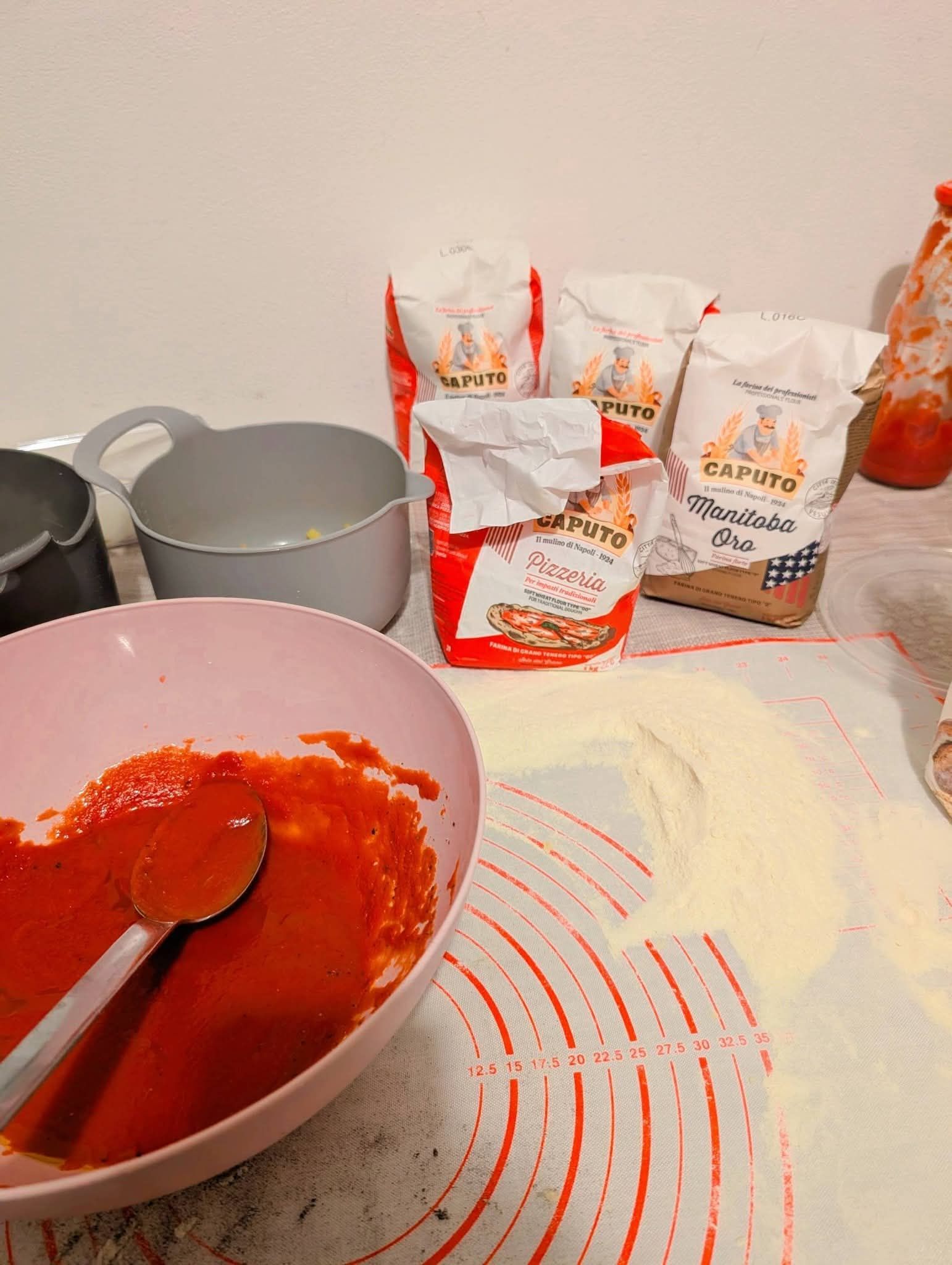 Una ciotola rosa di salsa di pomodoro rossa, un tappetino da forno in silicone con della farina e diversi sacchetti di farina sul piano della cucina.
