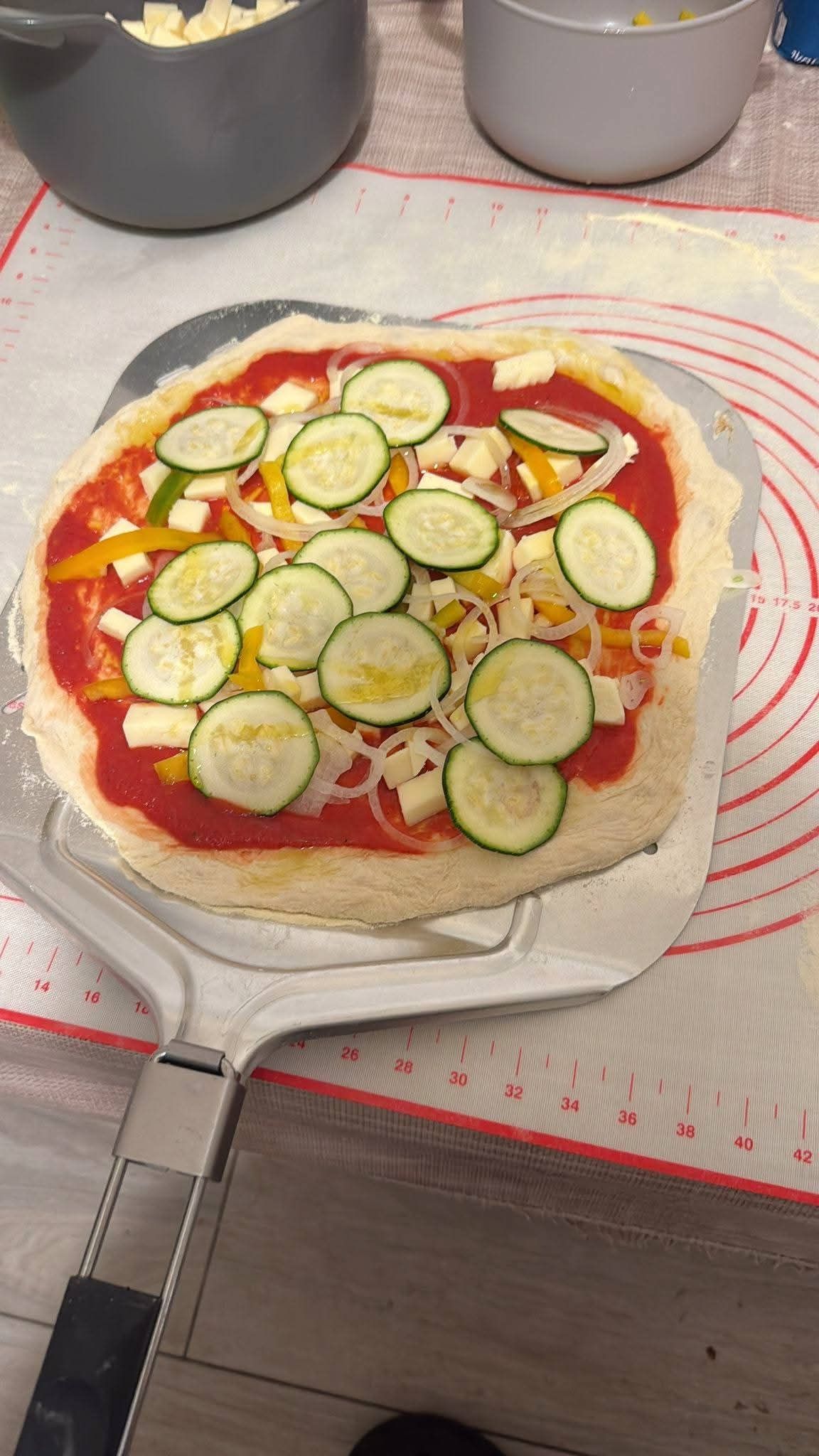 Una pizza condita con salsa di pomodoro, pezzi di mozzarella, zucchine a fette e peperoni è adagiata su una pala per pizza in metallo.