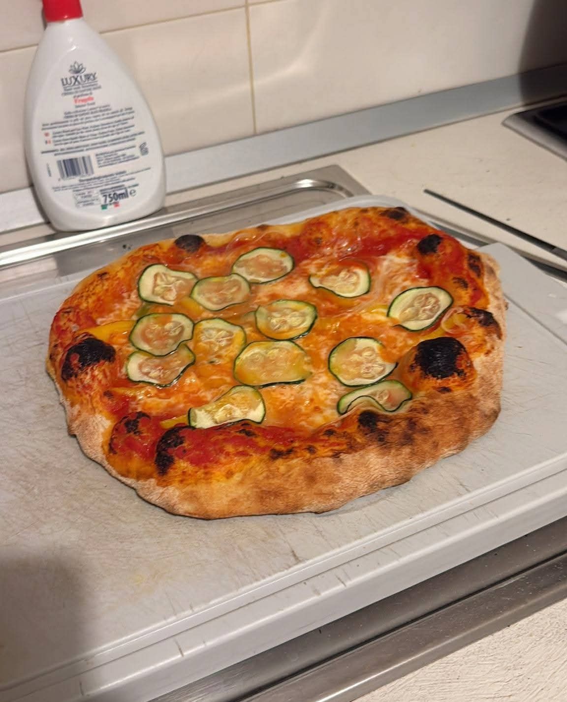 Una pizza fatta in casa condita con sottili fette di zucchine su un tagliere bianco, accanto a una bottiglia di detersivo per piatti.