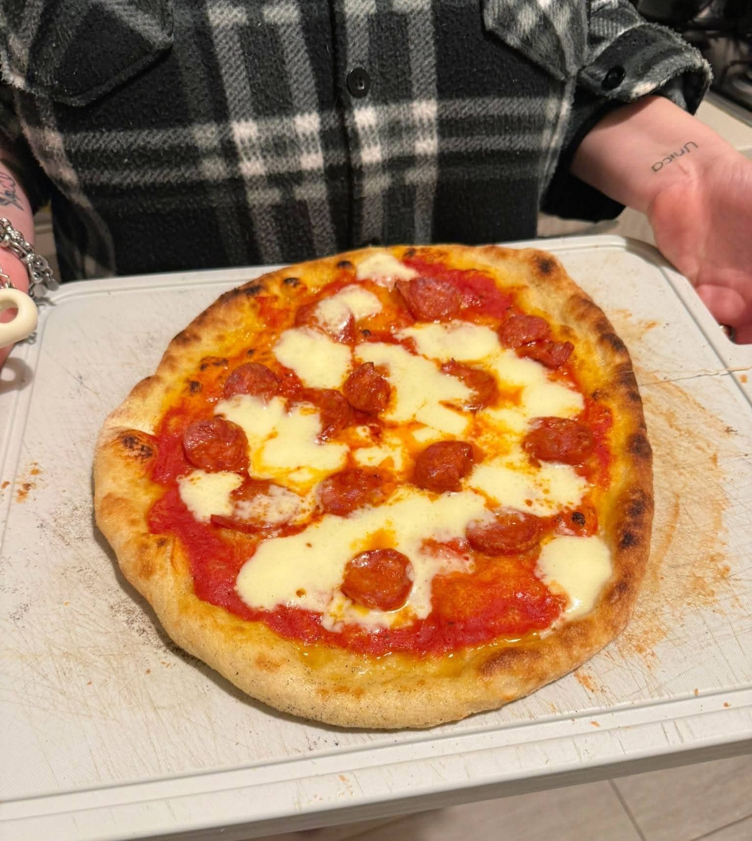 Una pizza fatta in casa con peperoni e mozzarella su un tagliere bianco, tenuta in mano da una persona che indossa una camicia di flanella a quadri.