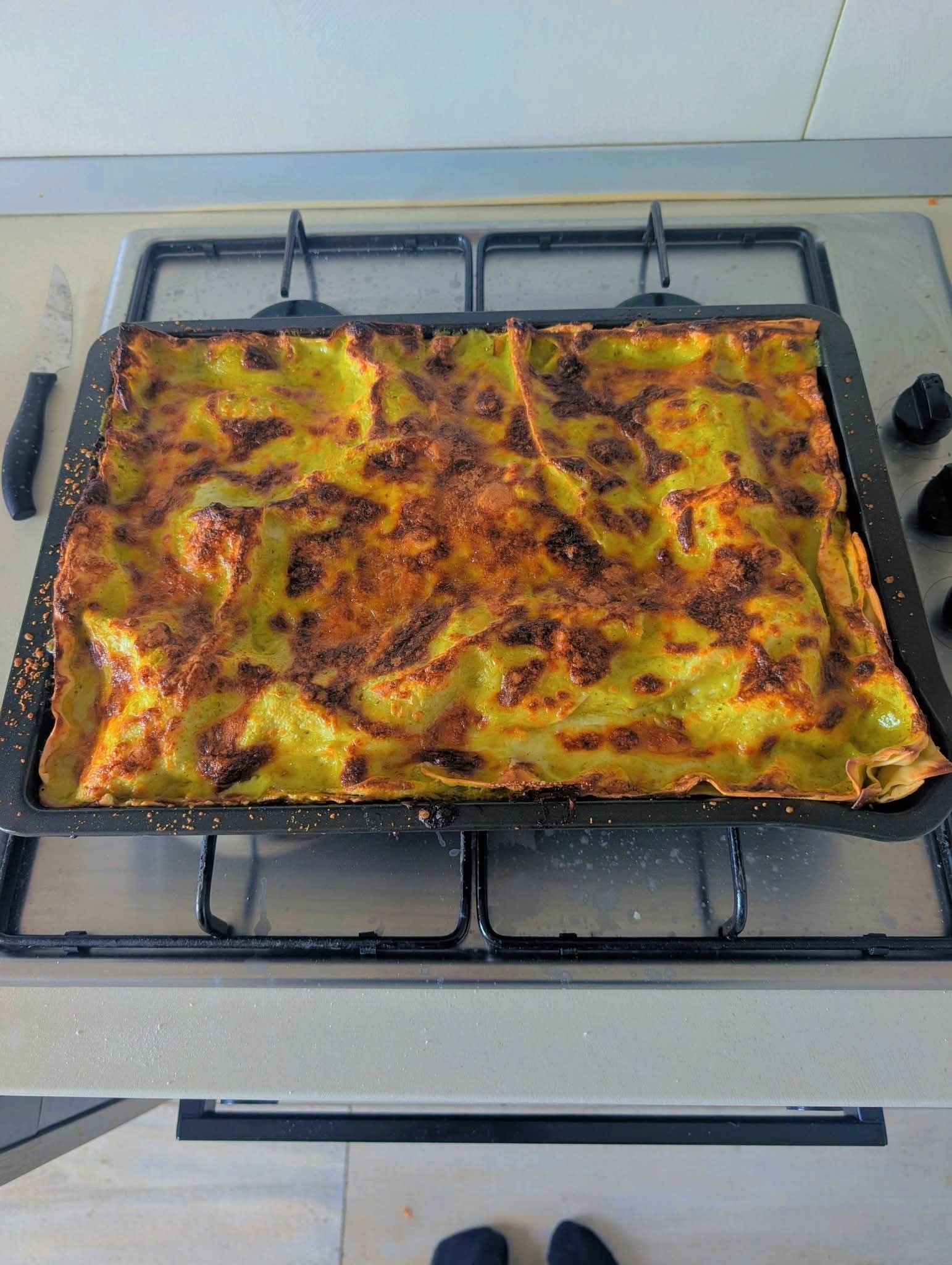 Una lasagna dorata cotta al forno è adagiata in una teglia rettangolare nera su un piano cottura in acciaio inossidabile.