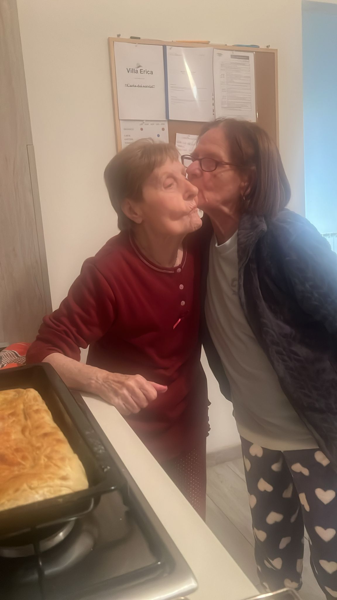 Due persone in cucina, una con una camicia rossa e l'altra con una giacca scura, si scambiano un bacio sulla guancia.