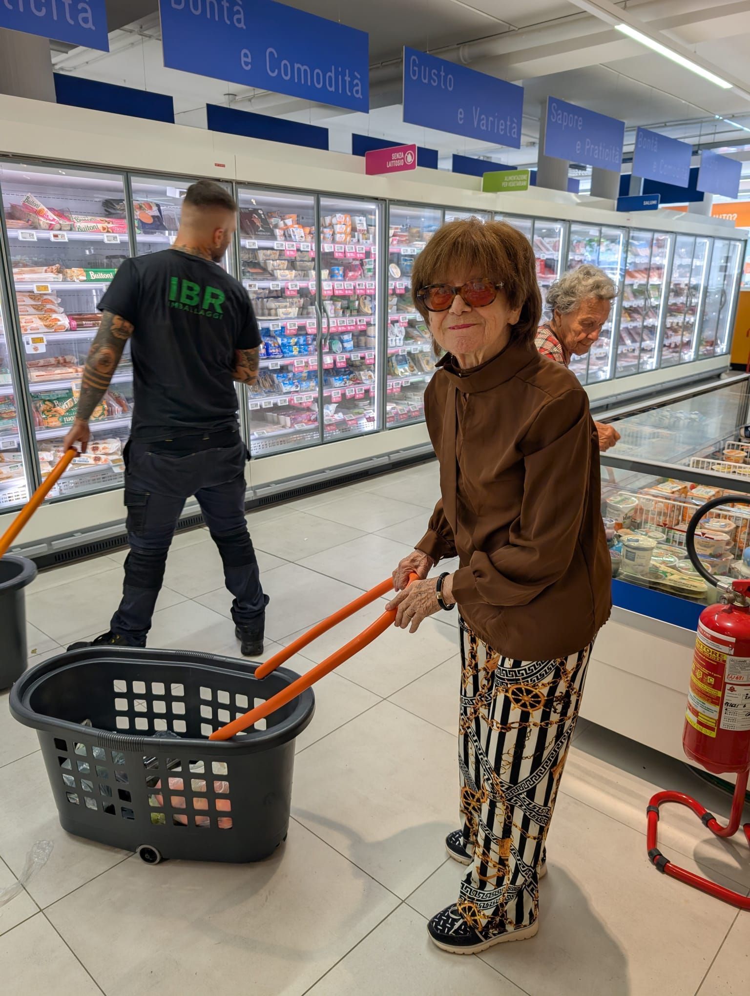 Una persona trascina un carrello in una corsia di un supermercato mentre un addetto rifornisce un banco frigo nelle vicinanze.