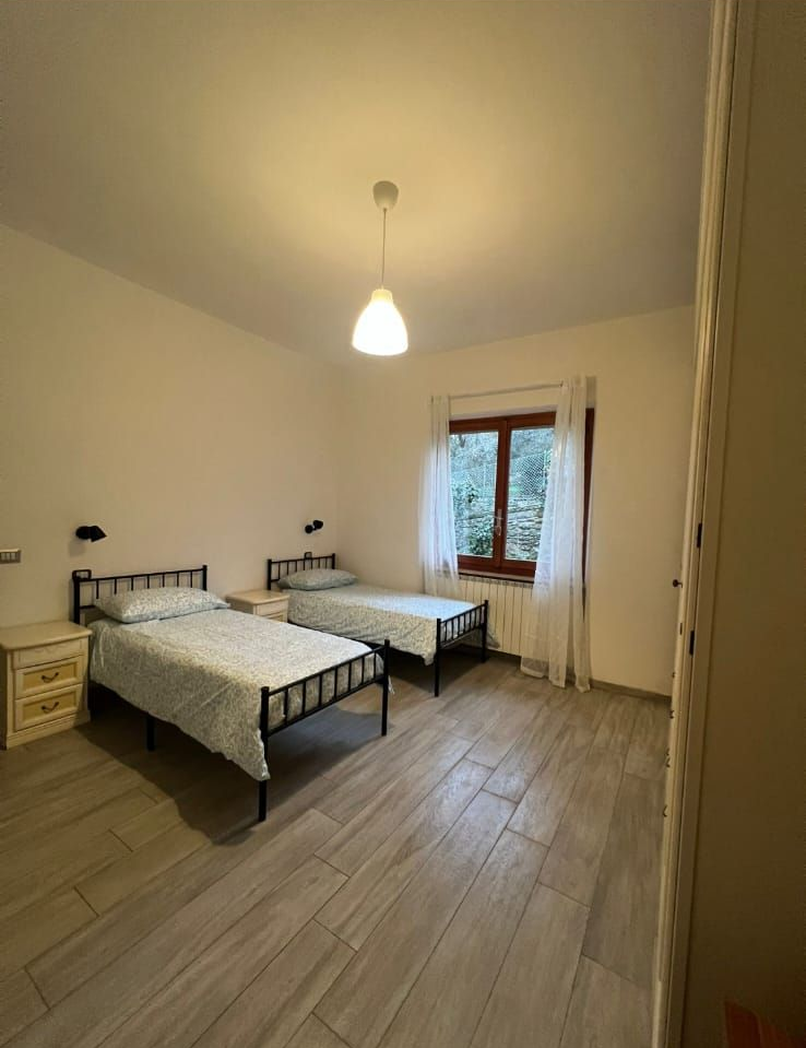 Una camera da letto luminosa con due letti singoli con struttura in ferro, pavimento in legno chiaro, lampada a sospensione e finestra con tende.