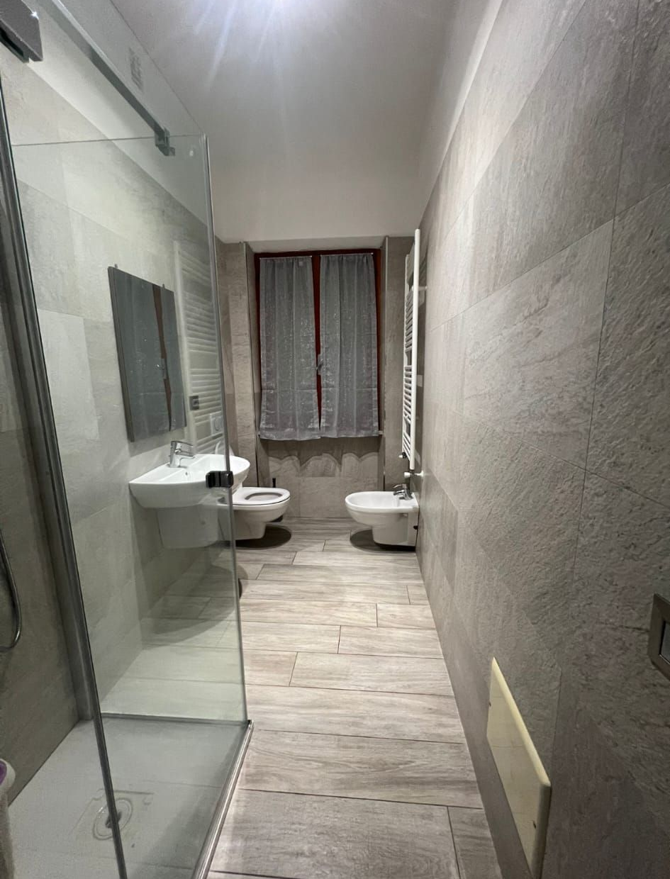 Un bagno moderno e stretto con box doccia in vetro, lavabo sospeso, WC e bidet, caratterizzato da pareti rivestite con piastrelle grigie.