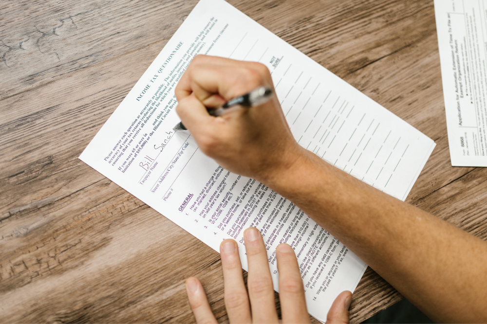 A person is signing a document with a pen on a wooden table.