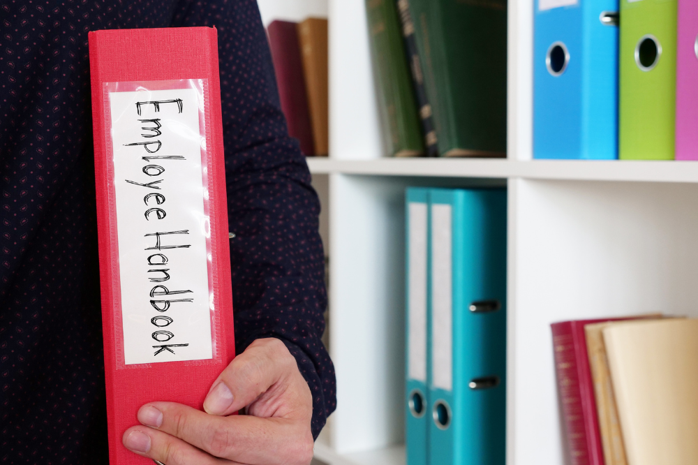 A person is holding an employee handbook in front of a bookshelf.