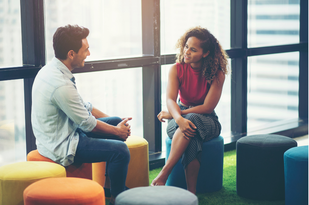 A man and a woman are sitting on colorful ottomans talking to each other.