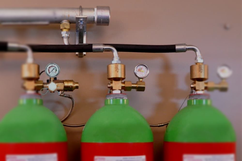 Three green and red gas cylinders are connected to a pipe on a wall.