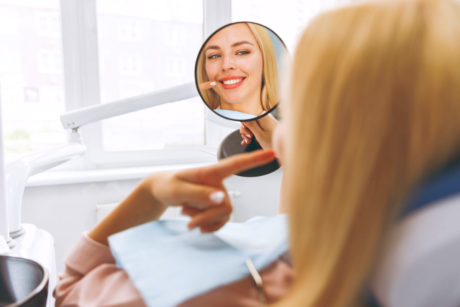 Woman in dentist chair looking at teeth in mirror; pointing to smile.