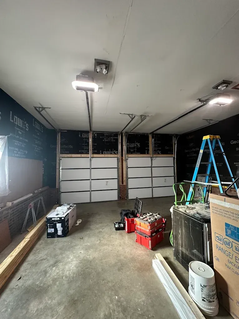 Garage under renovation with open door, ladder, tools, and construction materials scattered on the floor