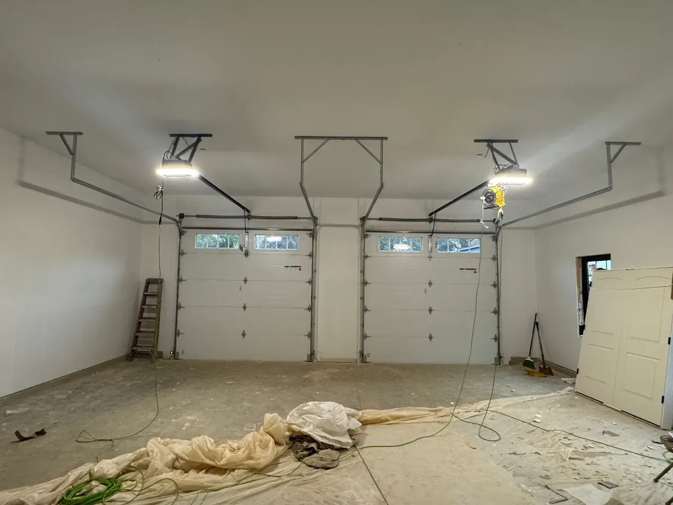 Empty white garage under construction with two overhead doors, concrete floor, and ladders against the wall