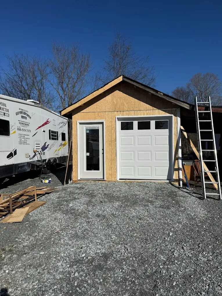 Small unfinished garage with white door and side entry, gravel driveway, and ladder beside it under a clear blue sky