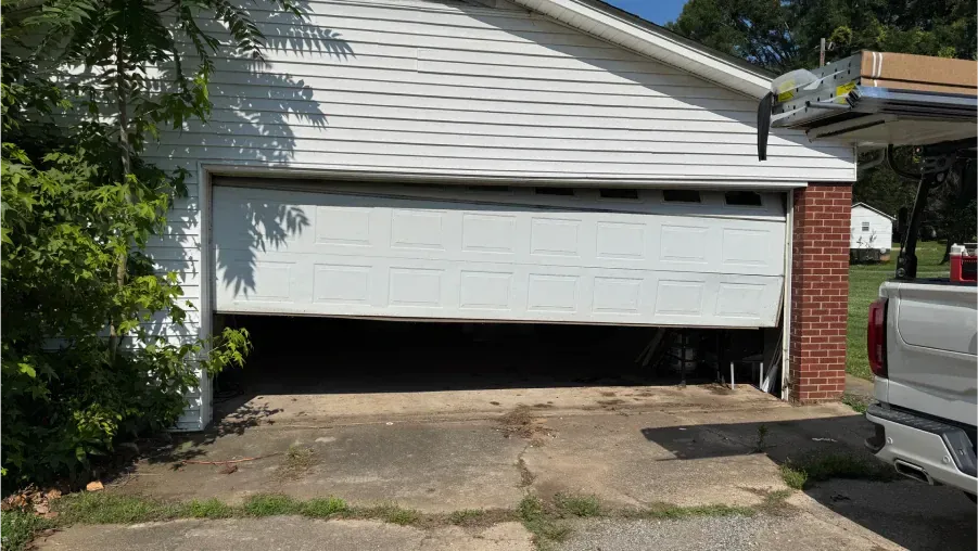 White garage door partly open on a house driveway, with a truck parked beside it and a brick pillar.