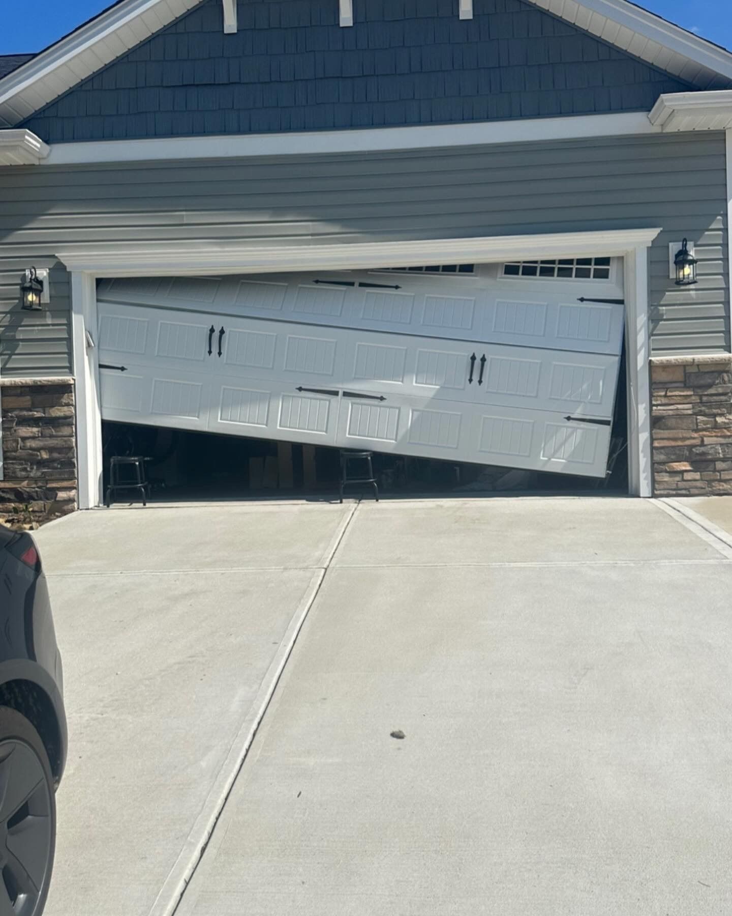 Tilted garage door hanging open on a house, with daylight visible inside and a driveway in front.